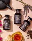 A flat lay photo showcases three glass bottles of The Goddess of Earl Set by Magic Hour, a goddess-themed Earl Grey tea collection. The bottles are beautifully displayed alongside a teapot, a filled teacup, loose tea leaves, the Gold Luxe Loose Leaf Tea Strainer, fresh lavender, and vibrant pink orchids on a beige surface.