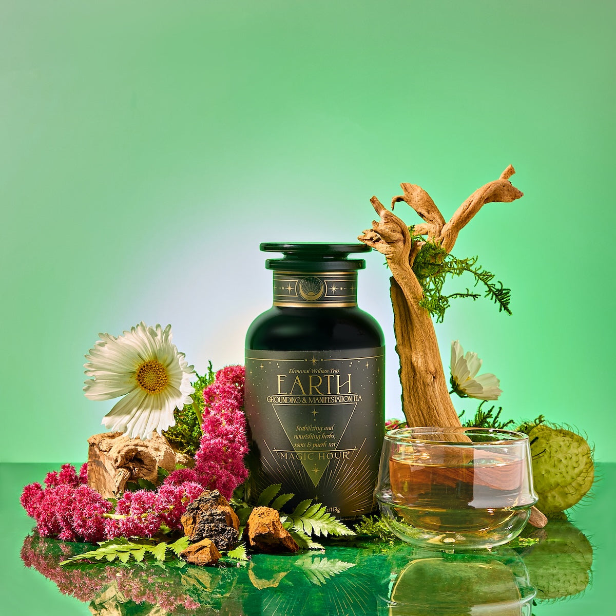 A dark green bottle labeled "Earth: Stabilizing and Nourishing Herbs, Roots, and Puerh tea" sits with flowers, bark, moss, a glass teacup of grounding tea, and a branch on a glossy surface against a green background for immune support.