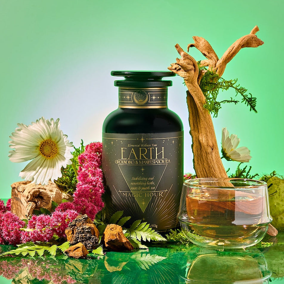 A dark green bottle labeled "Earth: Stabilizing and Nourishing Herbs, Roots, and Puerh tea" sits with flowers, bark, moss, a glass teacup of grounding tea, and a branch on a glossy surface against a green background for immune support.