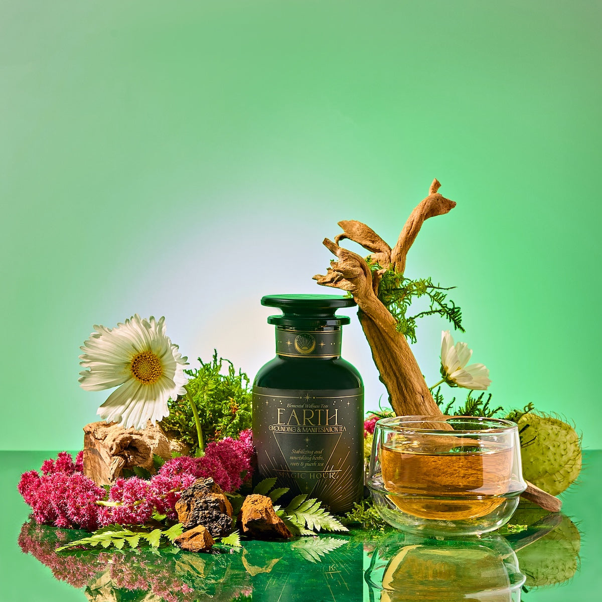 A dark green "Earth: Stabilizing and Nourishing Herbs, Roots, and Puerh tea" bottle sits among flowers, moss, wood, and herbs. Next to it is a clear cup of golden tea against a fresh green gradient background.