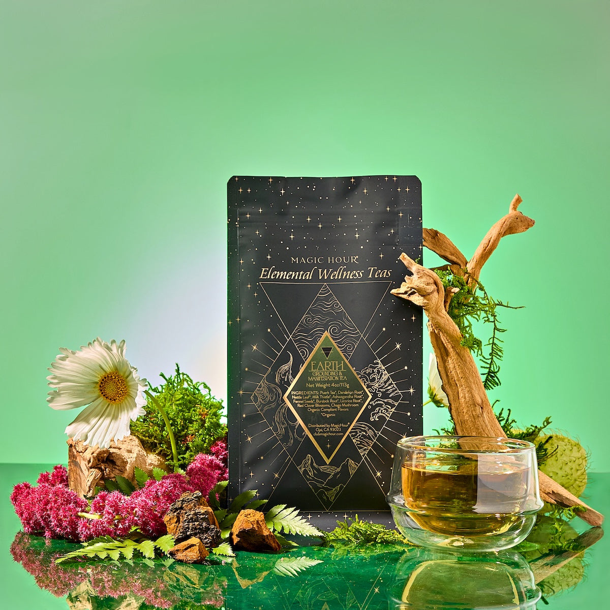 A black and gold tea package labeled "Earth: Stabilizing and Nourishing Herbs, Roots, and Puerh tea" rests among flowers, moss, driftwood, and a glass cup of grounding tea on a green gradient background.