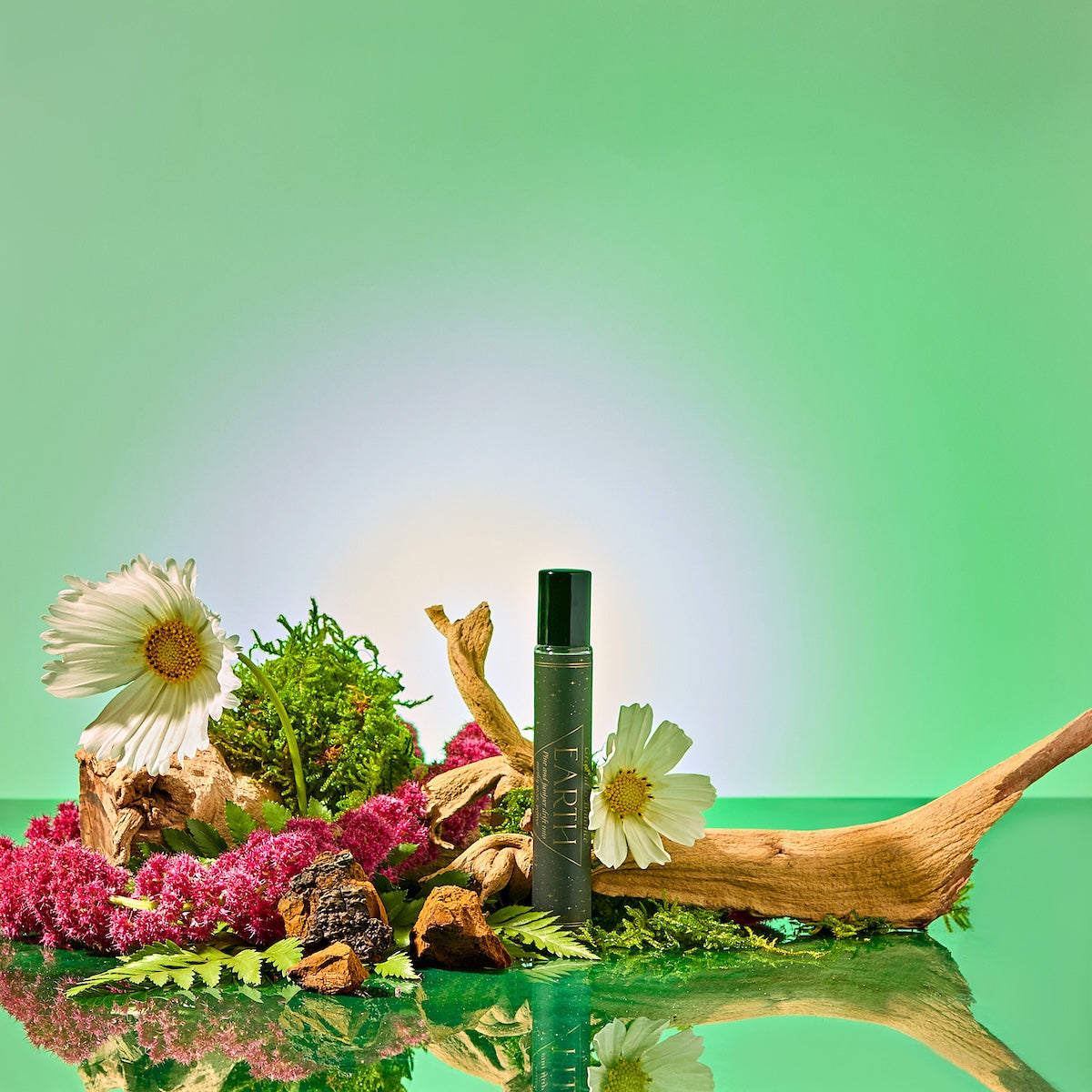 A black Earth Essential Oil Perfume bottle sits amid moss, pink and white flowers, ferns, bark, and driftwood on a reflective surface against a vibrant green gradient—a captivating botanical elixir inspired by nature.