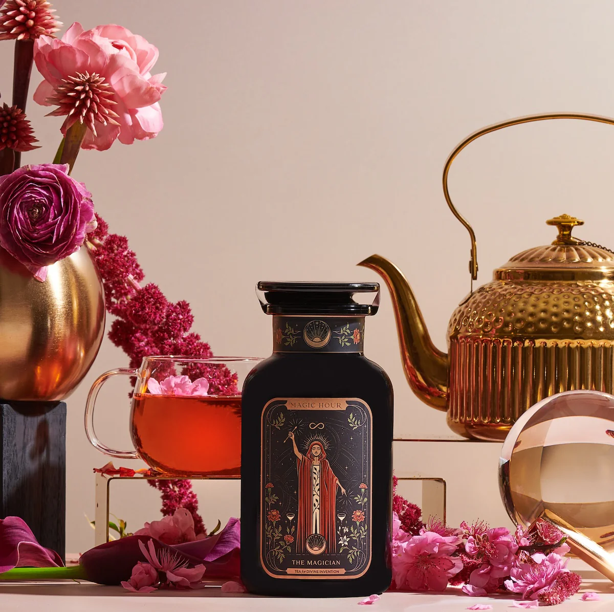 An arrangement of elegant tea-making items includes a tin labeled "The Magician" tea, a glass teacup filled with tea, various pink and red flowers, a gold teapot, and a crystal ball. The scene is set against a neutral backdrop, creating a sophisticated aesthetic.