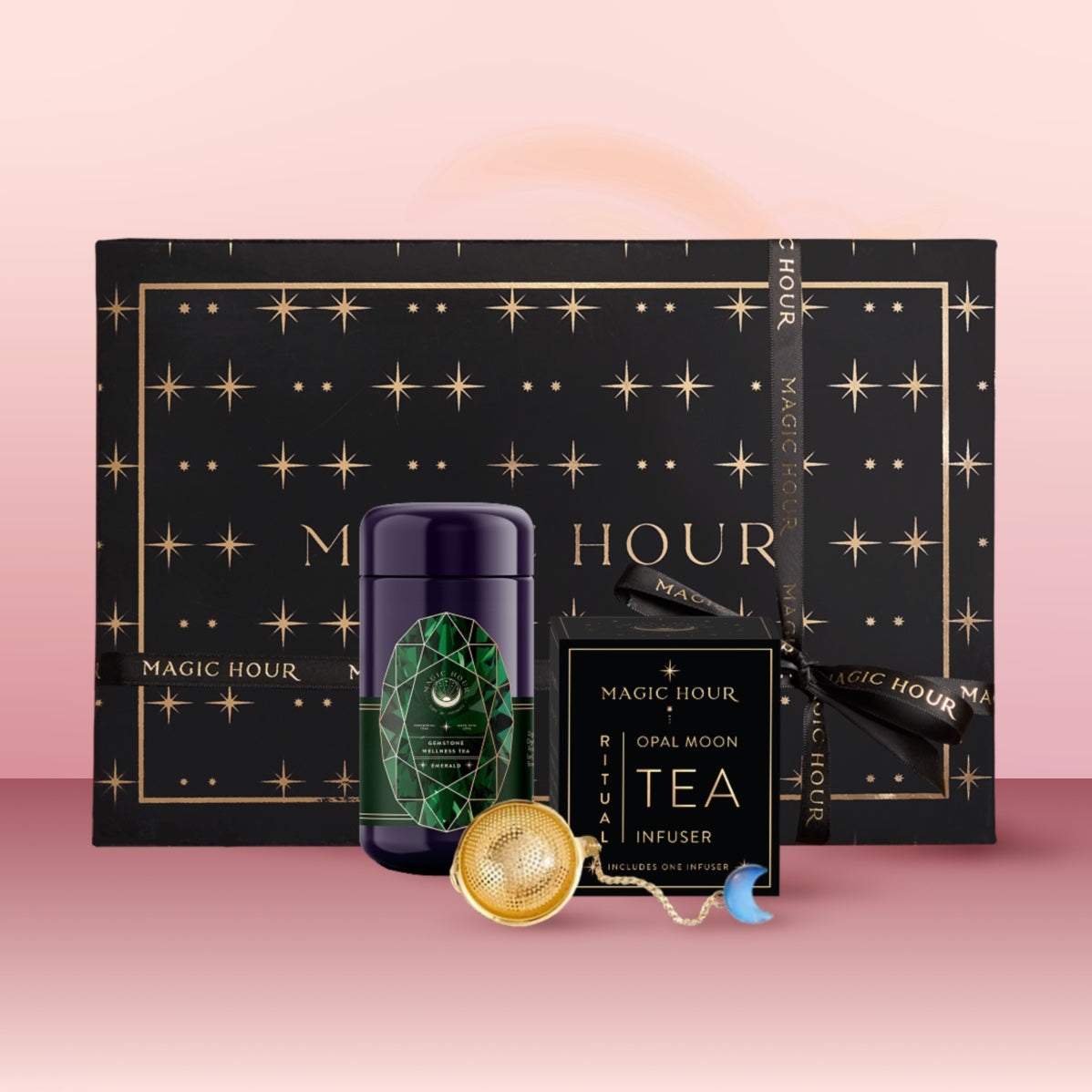 A black and gold star-patterned "Magic Hour" gift box sits behind a green tea canister, a tea infuser, and an "Opal Moon Tea Infuser" box on a pink surface—part of the Emerald May Birthstone Mini Gift Set.