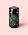 A black cylindrical container of Emerald - Cherry Blossom Pineapple Cream Green Tea, an organic Japanese green tea loose leaf wellness tea, featuring a green geometric label and set against a pale pink background.