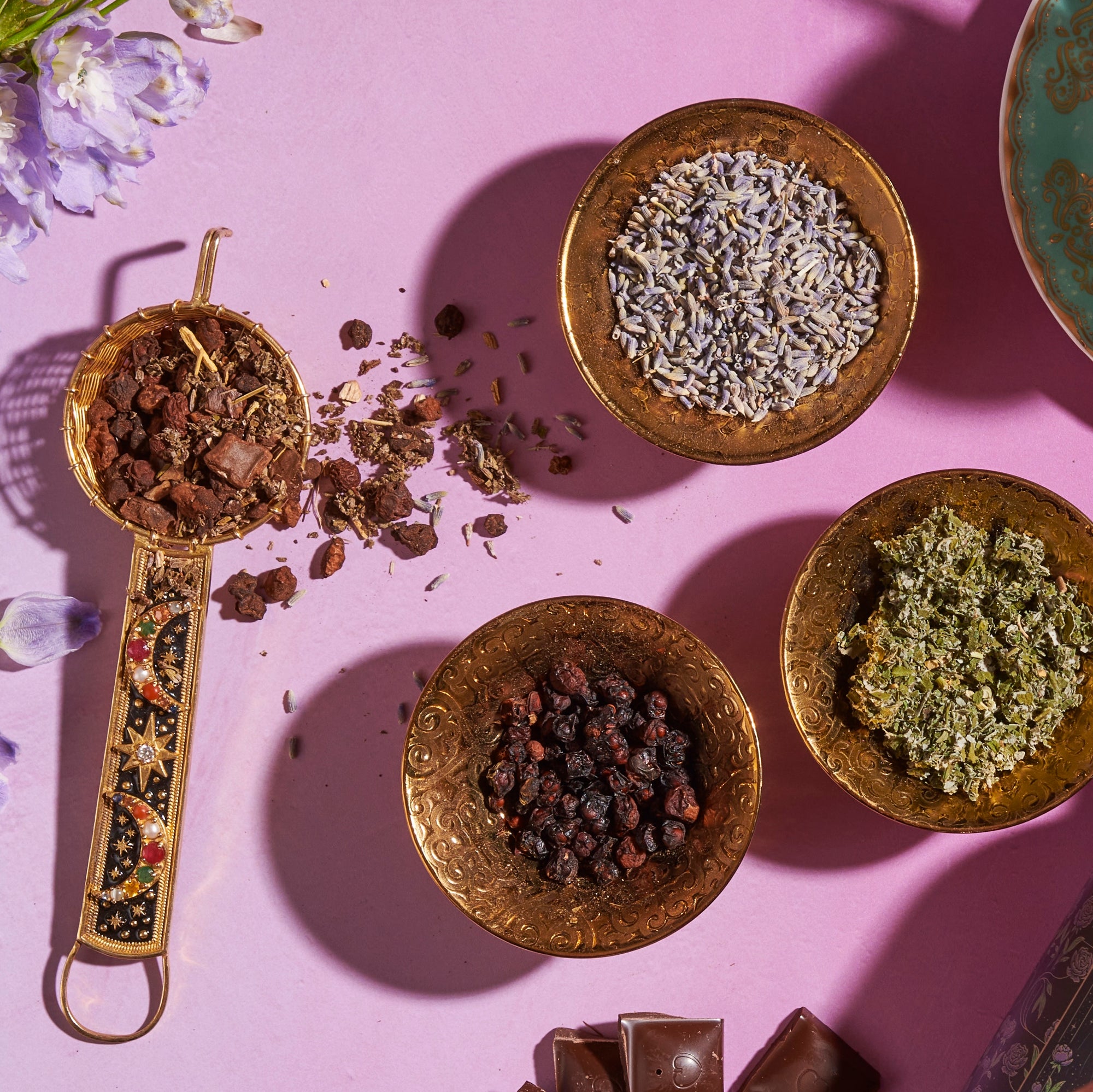 An overhead view of four ornate brass bowls on a pink surface. Each bowl contains different organic herbs and spices. The bowls are arranged neatly, casting shadows around them. Vibrant flowers and chocolate pieces surround the bowls, adding colorful touches reminiscent of a Magic Hour's The Empress: Lavender Currant Shatavari Cocoa Tea of Nurturing Creativity setup.