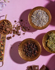 An overhead view of four ornate brass bowls on a pink surface. Each bowl contains different organic herbs and spices. The bowls are arranged neatly, casting shadows around them. Vibrant flowers and chocolate pieces surround the bowls, adding colorful touches reminiscent of a Magic Hour's The Empress: Lavender Currant Shatavari Cocoa Tea of Nurturing Creativity setup.