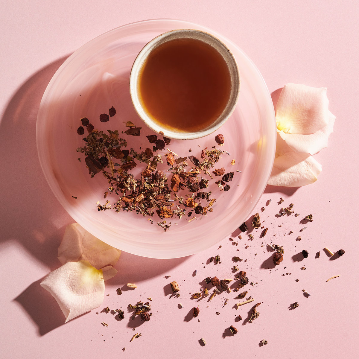 A delicate tea setup featuring a cup filled with Magic Hour's The Empress: Lavender Currant Shatavari Cocoa Tea of Nurturing Creativity on a pink saucer, surrounded by dried tea leaves and pink rose petals, all placed on a pink background. The arrangement has a soft and calming aesthetic.