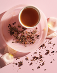 A delicate tea setup featuring a cup filled with Magic Hour's The Empress: Lavender Currant Shatavari Cocoa Tea of Nurturing Creativity on a pink saucer, surrounded by dried tea leaves and pink rose petals, all placed on a pink background. The arrangement has a soft and calming aesthetic.