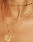 A close-up of a person wearing three layered necklaces: the top choker adorned with small green beads, the middle 18kt gold plate necklace showcasing the Enduring Individuality White Topaz Snake Necklace with a serpent symbol and white topaz detail, and the longest necklace displaying a round gold medallion with an intricate design.