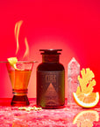 A cocktail with a lemon twist and flame sits beside a dark apothecary bottle labeled “Fire,” featuring crystal, ginger root, orange slice, and cardamom—showcasing adaptogens and metabolism-boosting herbs against a vibrant red backdrop.
