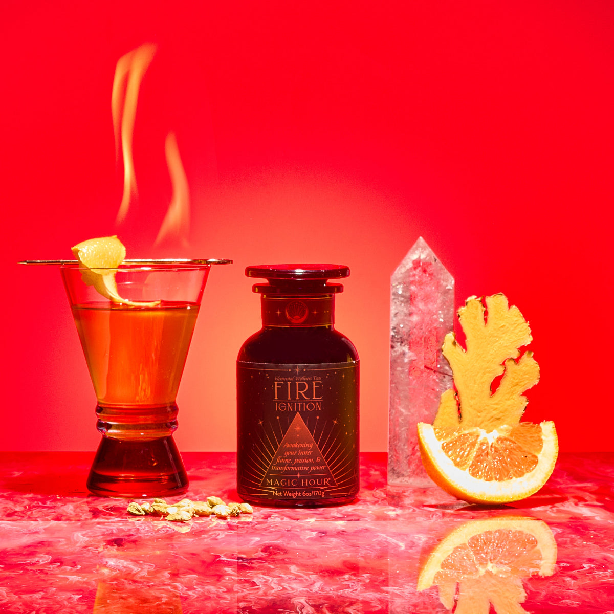 A vibrant red scene displays Fire—a cocktail with a lemon twist emitting flames, a dark "Fire" bottle, metabolism-supporting herbs, cardamom pods, ginger root, quartz crystal, Fire Tea, and orange on marble.