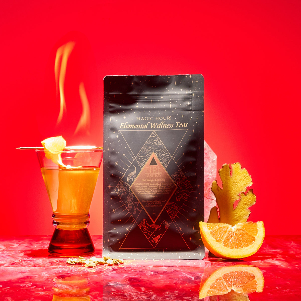 A glass of tea with lemon sits next to a black Magic Hour Fire package. Ginger, an orange slice, and a pink crystal rest on red marble, highlighting this metabolism-boosting herbal blend against a bold red background.