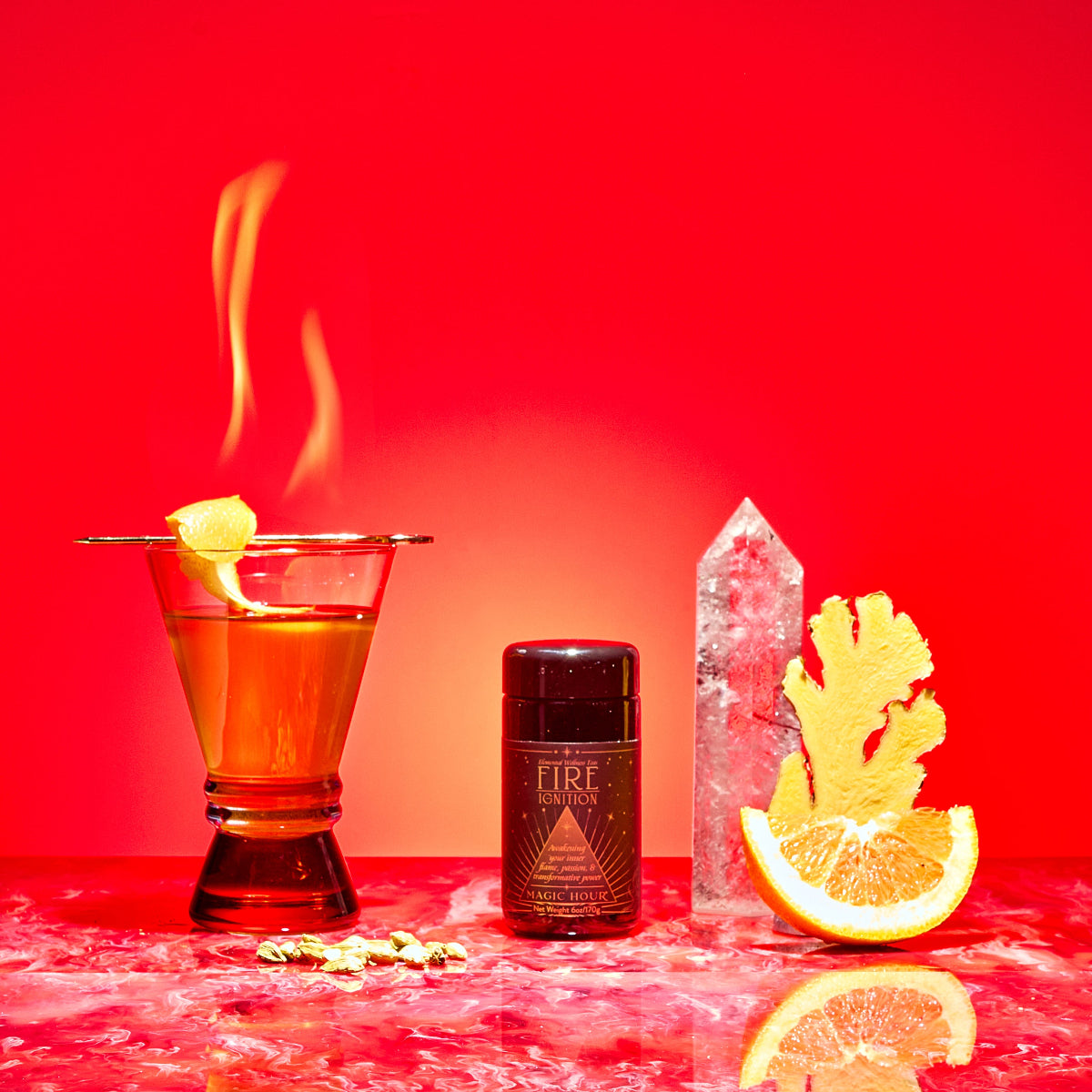 A cocktail with a flaming orange twist, a black "Fire" bottle, clear glass, ginger and orange slices sit on red marble with a vivid red backdrop—featuring Fire tea infused with metabolism-boosting herbs.