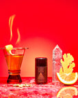 A cocktail with a flaming orange twist, a black "Fire" bottle, clear glass, ginger and orange slices sit on red marble with a vivid red backdrop—featuring Fire tea infused with metabolism-boosting herbs.