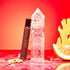 The Fire Essential Oil Perfume glass roller stands upright beside a clear quartz crystal point, cardamom pods, ginger root, and an orange slice on a red marbled surface with a red background.