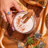 A hand holds a glass of creamy spiced chai, garnished with cinnamon sticks, resting on a wooden board. The scene is enriched by the Fall Flavors Tea Sampler Set, featuring an array of dried leaves and purple thistle flowers that enhance the warm autumn ambiance against the golden surface.