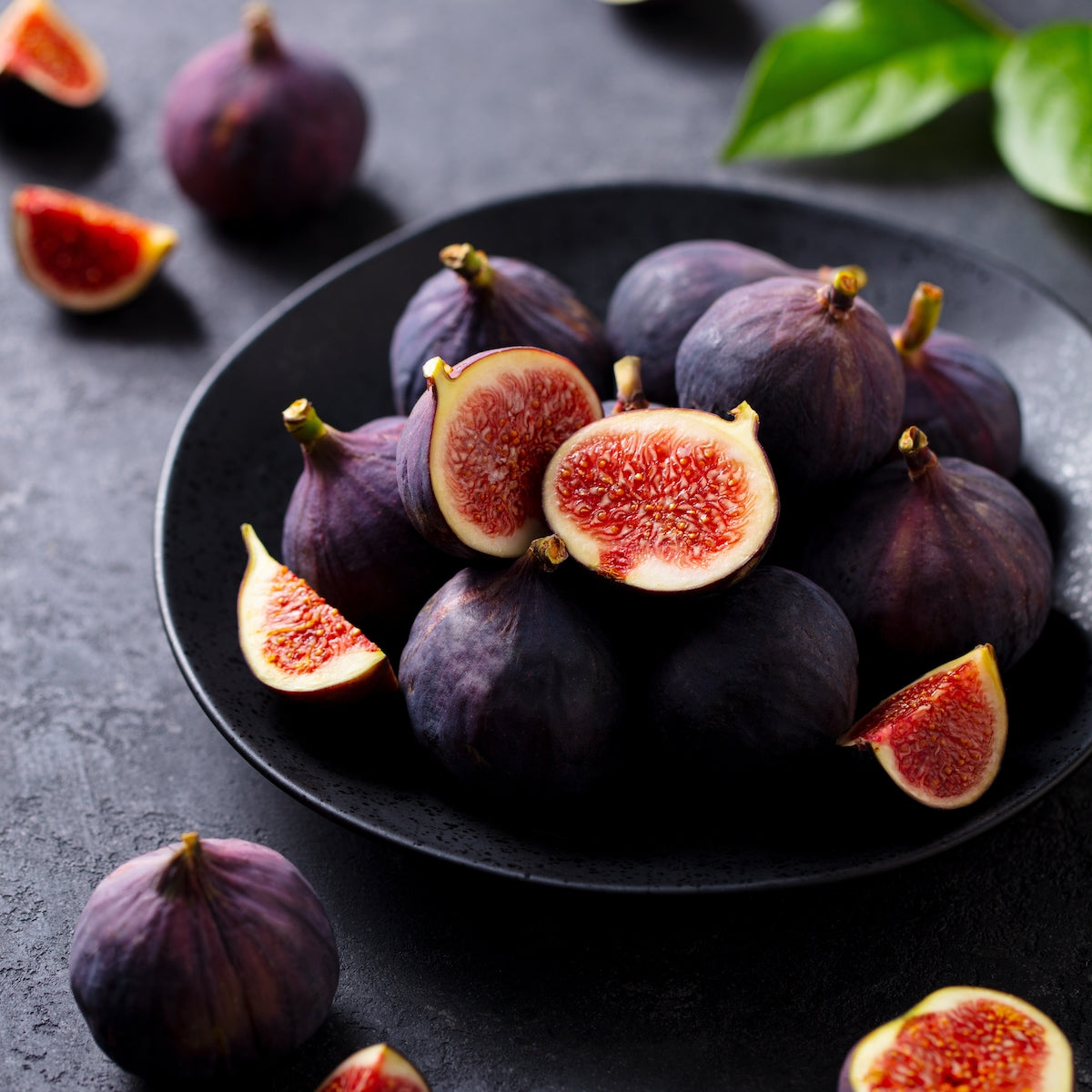 A black plate filled with whole and halved ripe figs is placed on a dark textured surface. Surrounding the plate are a few more whole and sliced figs. In the background, out of focus, are green leaves adding a touch of contrast to the dark scene.