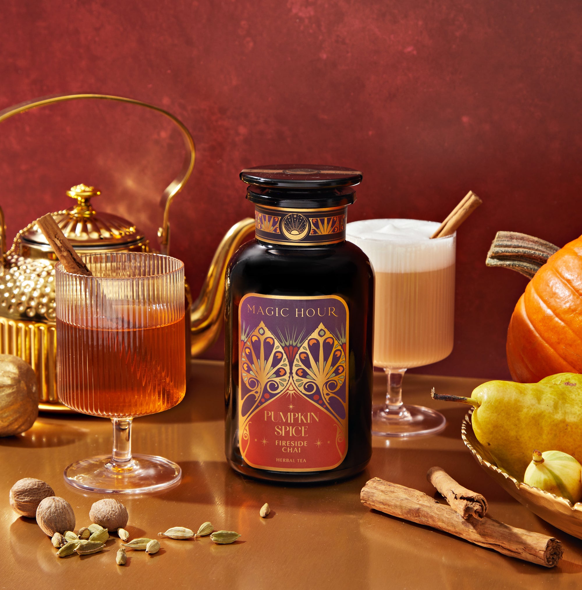 In a warm, reddish-brown setting, the "Pumpkin Spice Essentials Tea Trio Gift Set" features a black bottle adorned with its label. It is surrounded by two drinks garnished with cinnamon sticks, accompanied by a pumpkin, pear, and an assortment of spices. Fall flavors burst to life alongside a gold teapot and an arrangement of nutmegs in the background.