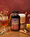 In a warm, reddish-brown setting, the "Pumpkin Spice Essentials Tea Trio Gift Set" features a black bottle adorned with its label. It is surrounded by two drinks garnished with cinnamon sticks, accompanied by a pumpkin, pear, and an assortment of spices. Fall flavors burst to life alongside a gold teapot and an arrangement of nutmegs in the background.