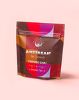 A colorful resealable pouch of Fireside Chai: Warming Rooibos Blend for Creating Connection—caffeine-free spiced herbal chai with Rooibos tea—in a red, orange, and purple geometric design on light pink. The packaging says "Sip Curiously.