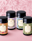 Four cylindrical tea canisters from the "Flower Powder: Matcha Bundle" by Magic Hour—featuring floral designs for Dragon Fruit, Blueberry Lavender Matcha, Elderberry Rose, and Strawberry Matcha—are arranged against a pink floral background and surface.