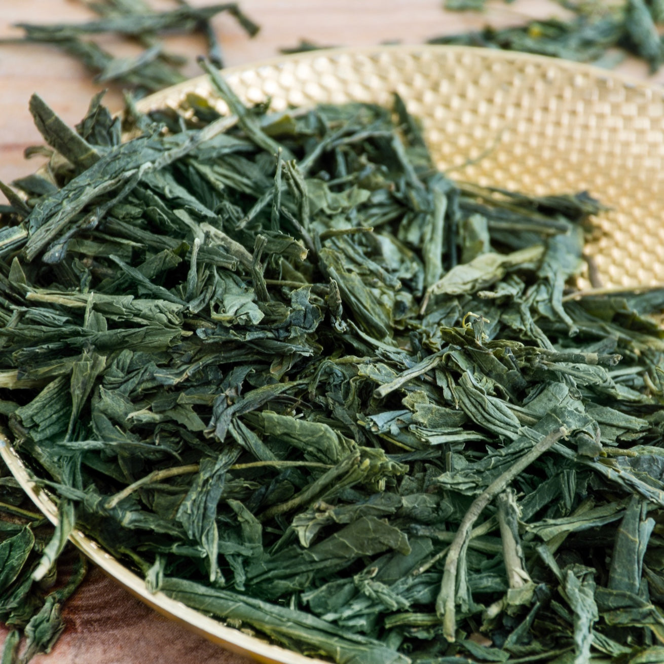 Loose dried Flower Fragrance Iccha Kariban Japanese green tea leaves on a textured golden plate, with some leaves scattered on a wooden surface in the background.
