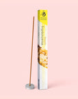 A box of Natural Incense : Frankincense, derived from desert-dwelling trees, stands upright next to a lit incense stick placed in a small round holder, all set against a pale pink background. The blissful fragrance gently fills the air, evoking tranquility and calm.