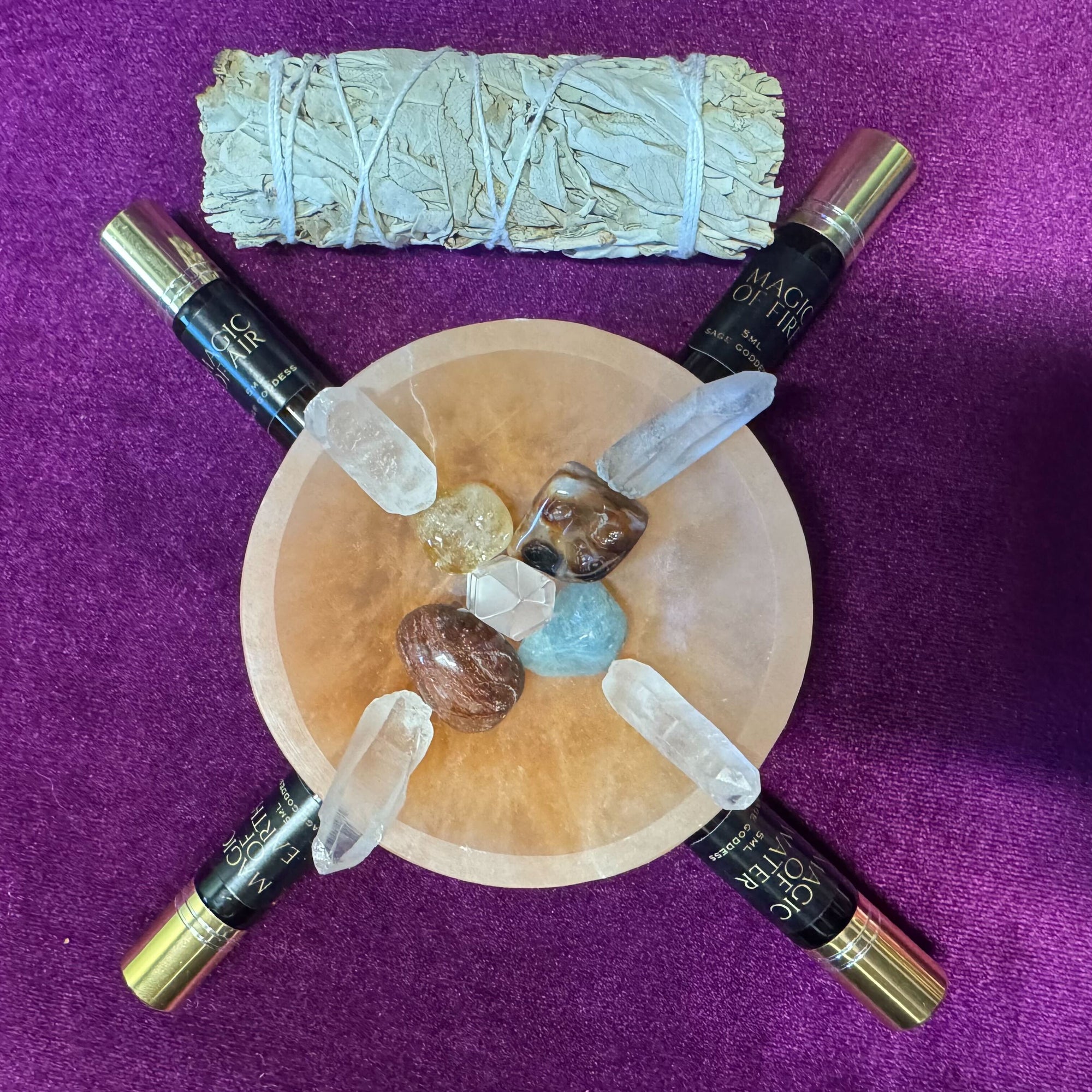 She Rises: A Transformational Year-End Ritual Retreat with Zhena Muzyka & Athena Perrakis features a pink salt crystal plate, assorted crystals, four labeled essential oil rollers, and dried sage—ideal for emotional healing and somatic practices.