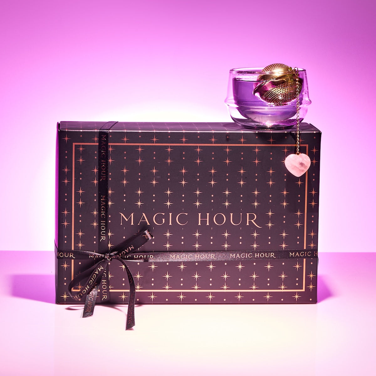 A black "Cycle Support Tea Subscription" gift box with a starry gold design and black ribbon, topped with a clear teacup, gold tea infuser, and heart charm—perfect for PMS relief—set on a purple-pink gradient background.