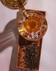 A Tea Discovery Club glass cup filled with tea rests on an ornate saucer atop a wooden tray, curated tea blends scattered nearby and a golden teapot pouring tea from above.