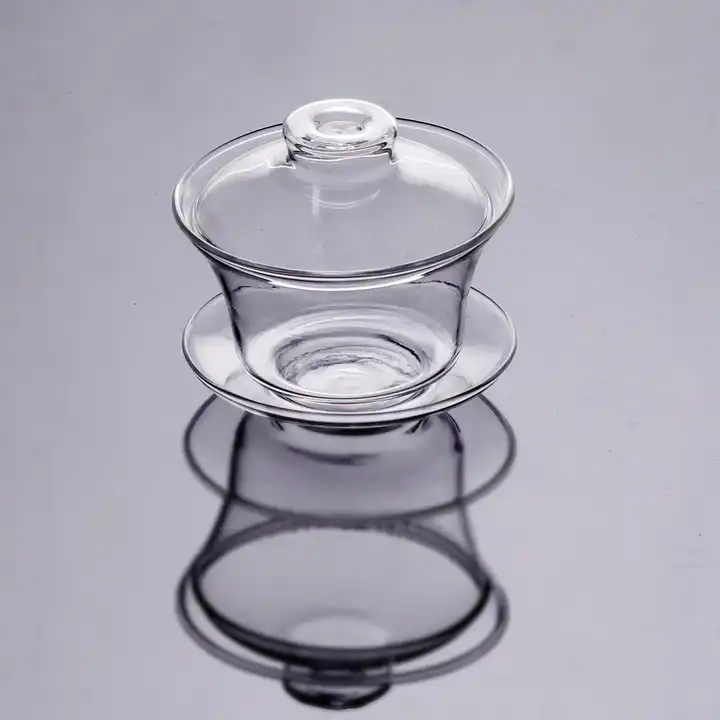 A Clear Glass Gaiwan, consisting of a tea cup, matching lid, and saucer, is elegantly displayed on a reflective surface, producing a mirrored effect. Its simple yet sophisticated design showcases smooth curves and a shiny finish.