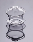 A Clear Glass Gaiwan, consisting of a tea cup, matching lid, and saucer, is elegantly displayed on a reflective surface, producing a mirrored effect. Its simple yet sophisticated design showcases smooth curves and a shiny finish.