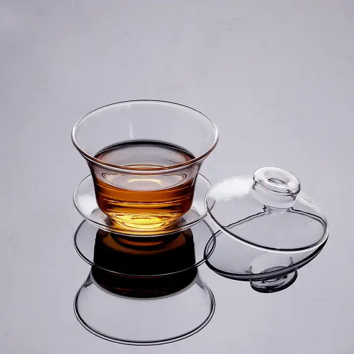 A Clear Glass Gaiwan, filled with amber-colored tea, rests on a saucer, with its matching glass lid placed beside it. The set is positioned on a smooth, reflective surface.