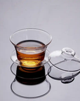 A Clear Glass Gaiwan, filled with amber-colored tea, rests on a saucer, with its matching glass lid placed beside it. The set is positioned on a smooth, reflective surface.