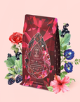 A red tea package adorned with a crystal design is surrounded by illustrated flowers, berries, and leaves on a pale pink background. The label reads “Gemstone Wellness Tea • Garnet: Violet Wine,” inviting you to experience this vibrant organic herbal potion—Garnet: Violet Wine Gemstone Wellness Tea—crafted for vitality and balance.