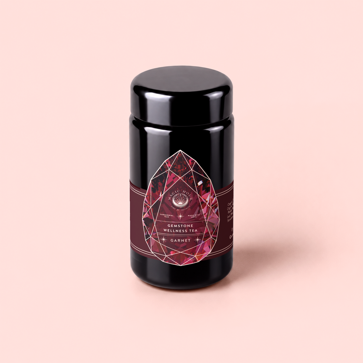 A black tea canister labeled “Garnet: Violet Wine Gemstone Wellness Tea,” featuring a red gemstone design on the front, is set against a pale pink background. Inspired by the energizing properties of garnet, this vibrant vitality tea is perfect for anyone seeking a confidence boost in every cup.