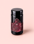 A black tea canister labeled “Garnet: Violet Wine Gemstone Wellness Tea,” featuring a red gemstone design on the front, is set against a pale pink background. Inspired by the energizing properties of garnet, this vibrant vitality tea is perfect for anyone seeking a confidence boost in every cup.