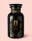 A black perfume bottle rests on a pale pink background, adorned with gold Gemini zodiac twins, stars, a crescent moon, and lilies on its label. The elegant design features the words “Gemini: Watermelon-Rose-Mulberry Pomegranate Tea for Beauty, Balance & Quenching Curiositeas,” while delicate accents of jasmine and organic rose petals evoke the fragrance’s inspiration.