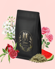 A black tea package labeled "Gemini: Watermelon-Rose-Mulberry Pomegranate Tea for Beauty, Balance & Quenching Curiositeas" is surrounded by rose petals, pomegranate, watermelon slice, Silver Needle White Tea, and loose tea leaves on a light pink background.