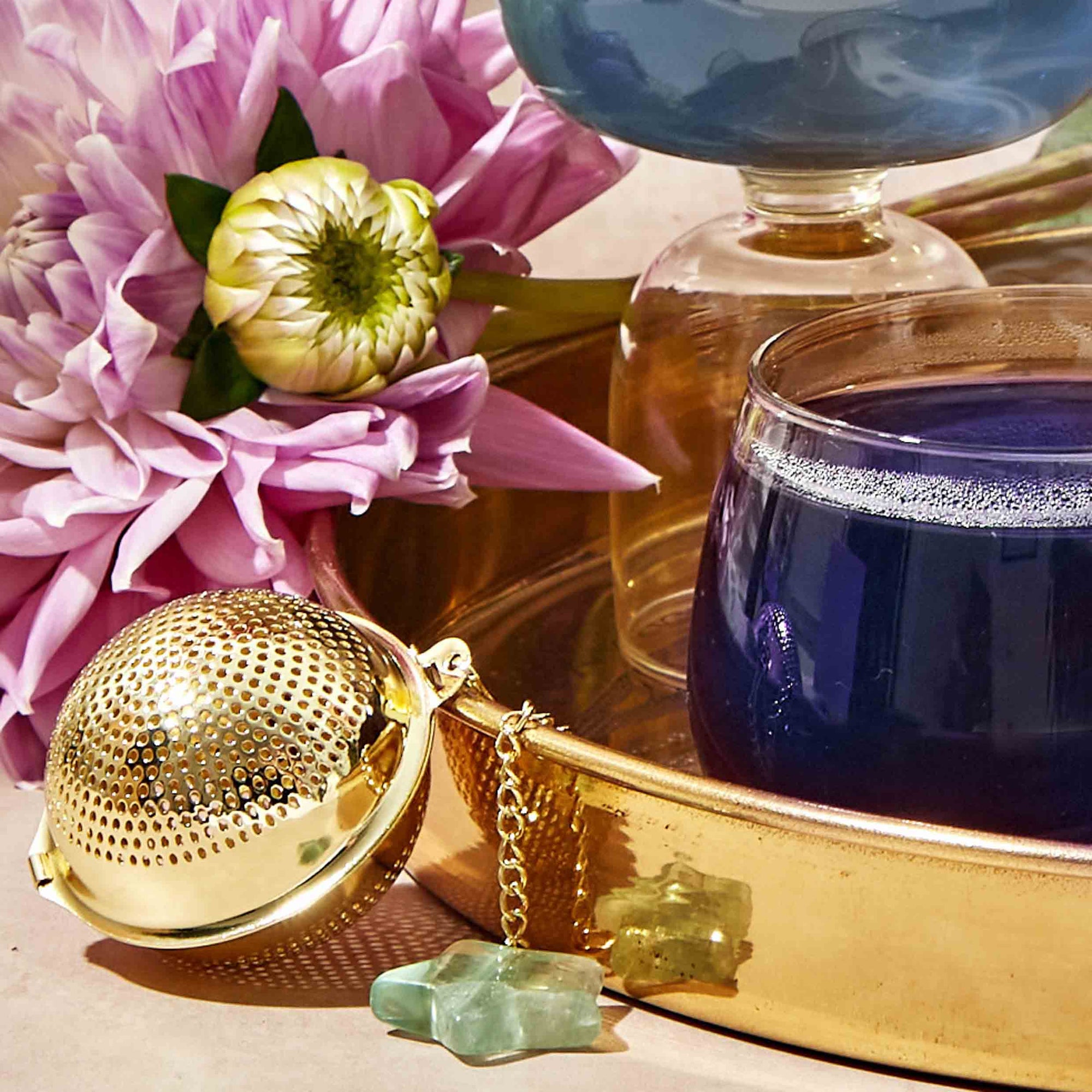 A gold Emerald Star Tea Strainer rests on a shiny gold tray alongside purple and white flowers and a glass of blue liquid. In the background, there is another blue drink in a glass, and two small stones are placed next to the tea strainer.