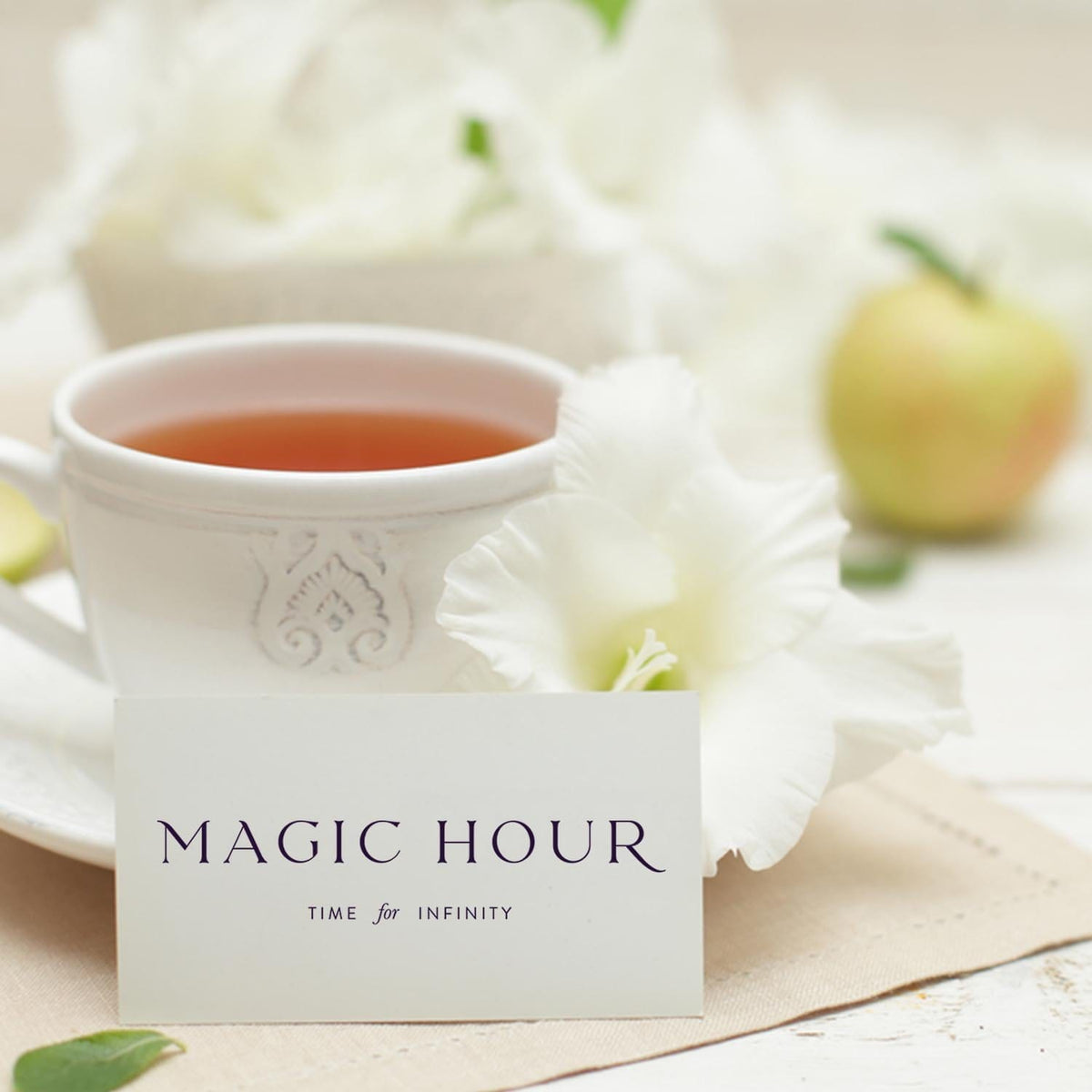 A white teacup with tea sits on a napkin beside a white flower and an apple. In front is a card labeled "Gift Card," hinting at the ideal present—a Gift Card. The softly blurred background adds to the charm.