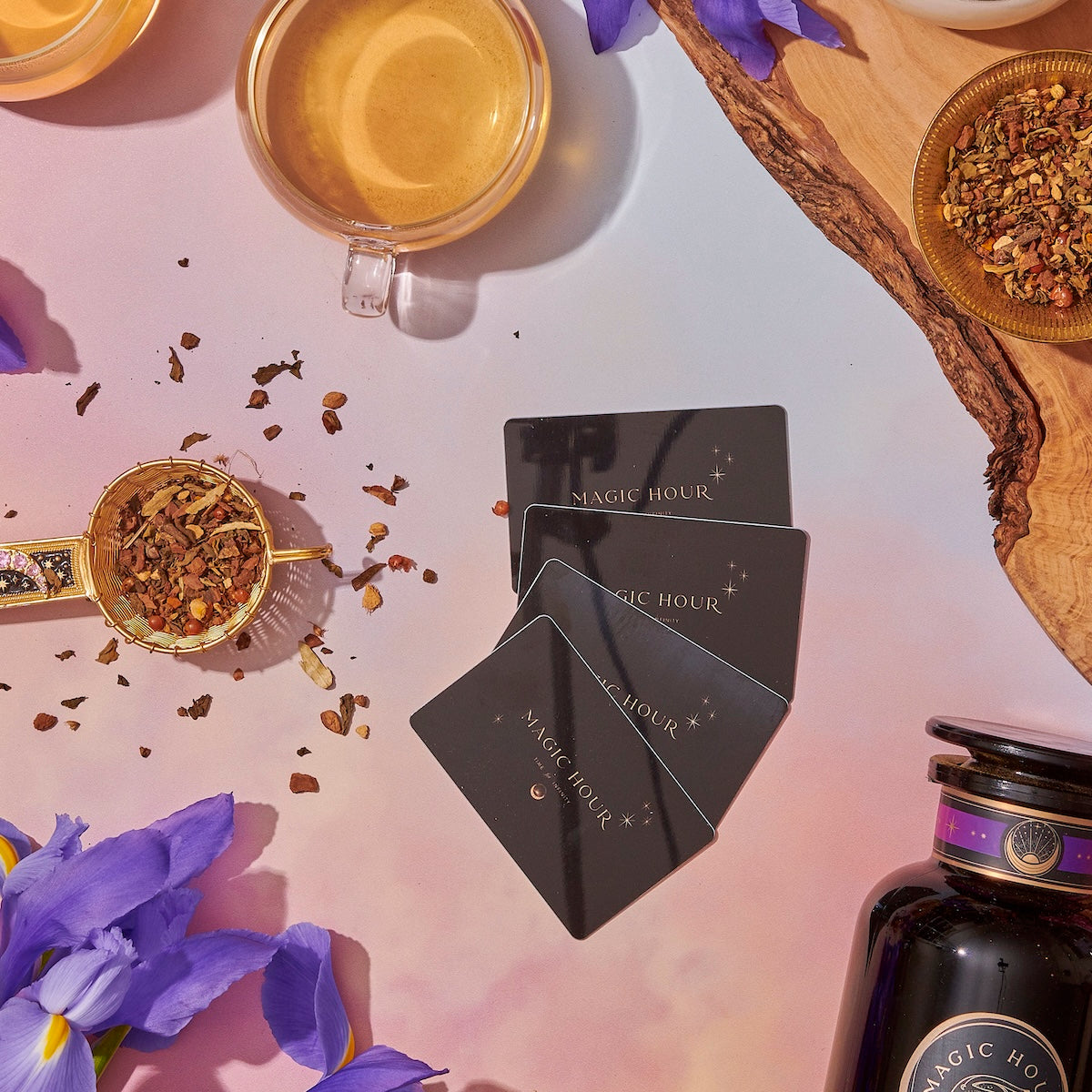 A pastel flat lay shows a cup of tea, gold strainer with loose leaves, purple flowers, dried herbs, a jar, and four black Club Magic Hour Gift Cards—perfect for emailing a Gift Card to someone special.