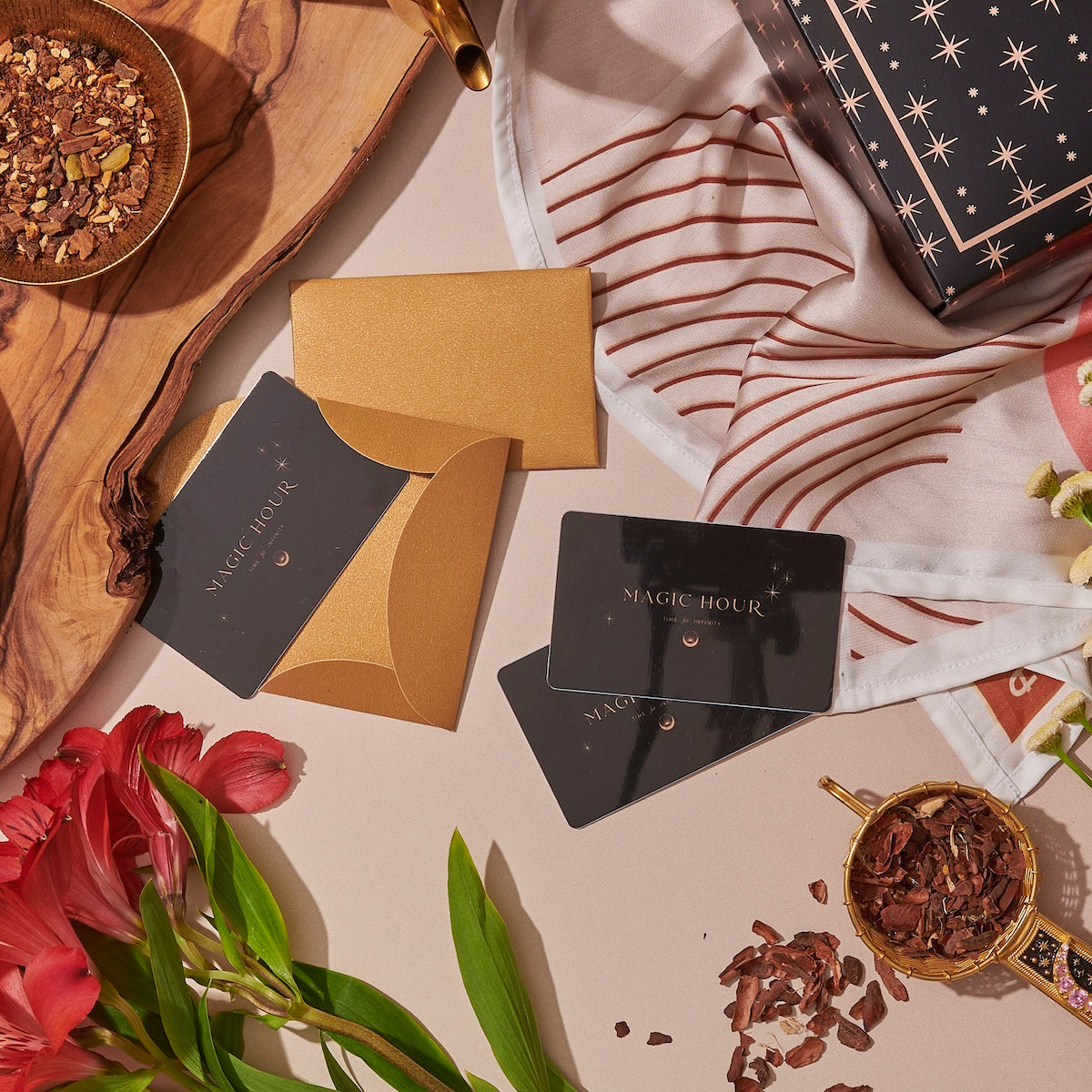 A flat lay of the Gift Card set includes a gold envelope with "Magic Hour" cards, a wrapped present adorned with stars, a Club Magic Hour gift card for redemption at checkout, a cup of loose tea, tea leaves arranged on a wooden board, and flowers in the picture. The aesthetic is warm and inviting with tones of gold, red, and brown.