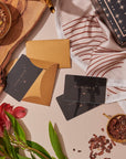 A flat lay of the Gift Card set includes a gold envelope with "Magic Hour" cards, a wrapped present adorned with stars, a Club Magic Hour gift card for redemption at checkout, a cup of loose tea, tea leaves arranged on a wooden board, and flowers in the picture. The aesthetic is warm and inviting with tones of gold, red, and brown.