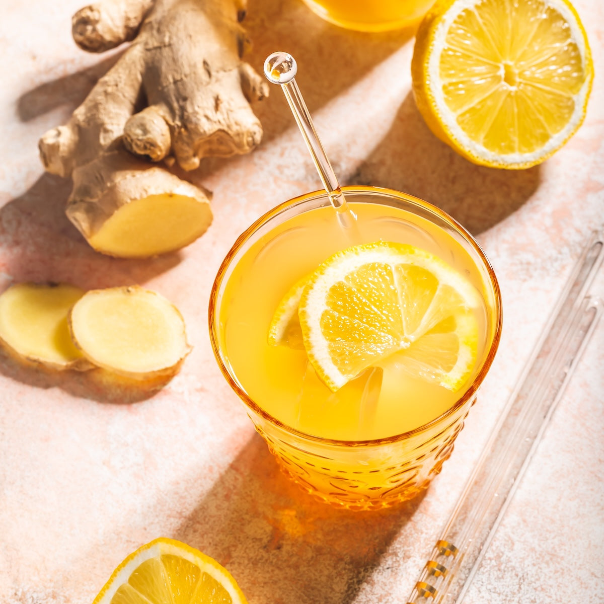 A glass of Ginger Lemon with lemon slices, surrounded by fresh ginger root, ginger slices, and lemon halves on a light surface. A glass straw is in the drink.