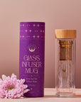 An Alibaba Glass Infuser Mug with a bamboo lid stands next to its purple cylindrical packaging labeled "Glass Infuser Mug for Loose Leaf Tea & Fruit Water." The detachable infuser and double-walled borosilicate glass ensure durability. A pale pink chrysanthemum flower is placed at the base of the packaging, all set against a neutral background.