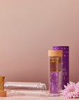 A clear glass infuser mug complete with a wooden lid and gold star accents, sits beside its star-adorned cylindrical purple packaging. The Glass Infuser Mug also boasts a detachable infuser inside. To the right of the mug, a pink chrysanthemum flower rests on the surface.