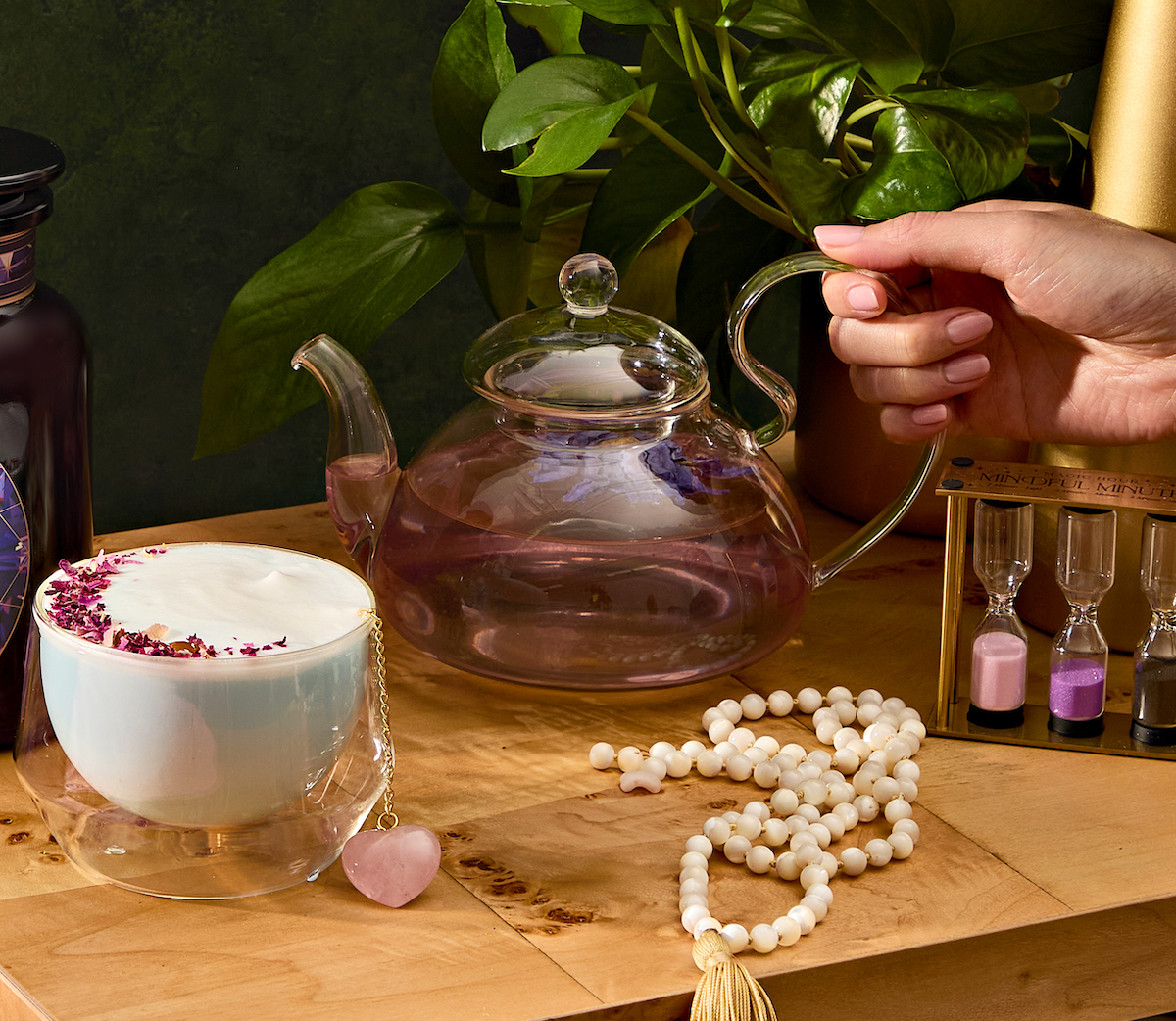 An Angel Glass Tea Pot, ideal for brewing 4 cups of loose leaf tea with mesmerizing purple liquid, is cradled in a hand. Nearby, a creamy topped beverage with dried flowers sits alongside a rosary and hourglasses nestled among green plants on the wooden table.