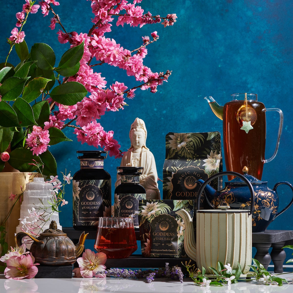 A colorful arrangement featuring a variety of organic tea products displayed on a table. The scene includes teapots, tea canisters, packets of Magic Hour&#39;s Goddess of Earl: Madagascar Vanilla Creme Tea for Soothing Delight &amp; Delicious Decadence, a glass of tea, and a glass pitcher of iced tea with a star-shaped tea infuser. Pink cherry blossoms and a Buddha statue adorn the background.